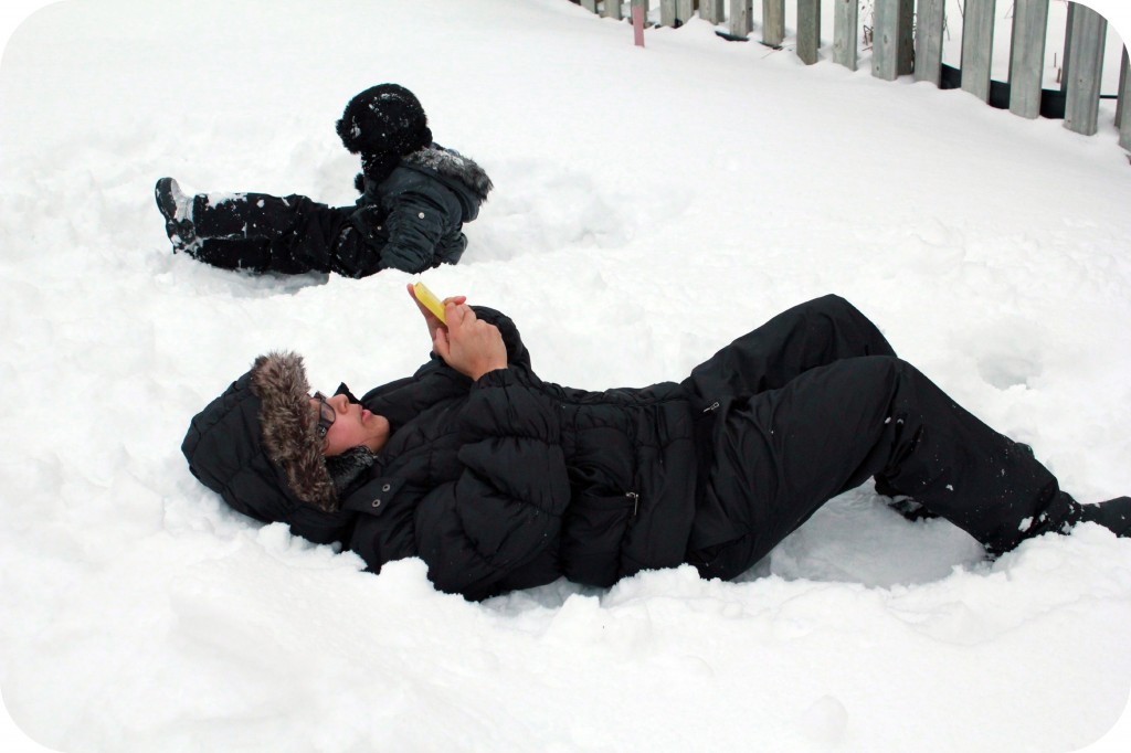 Snowmageddon. A little girl plays in the snow while mother tweets on her phone on the ground.