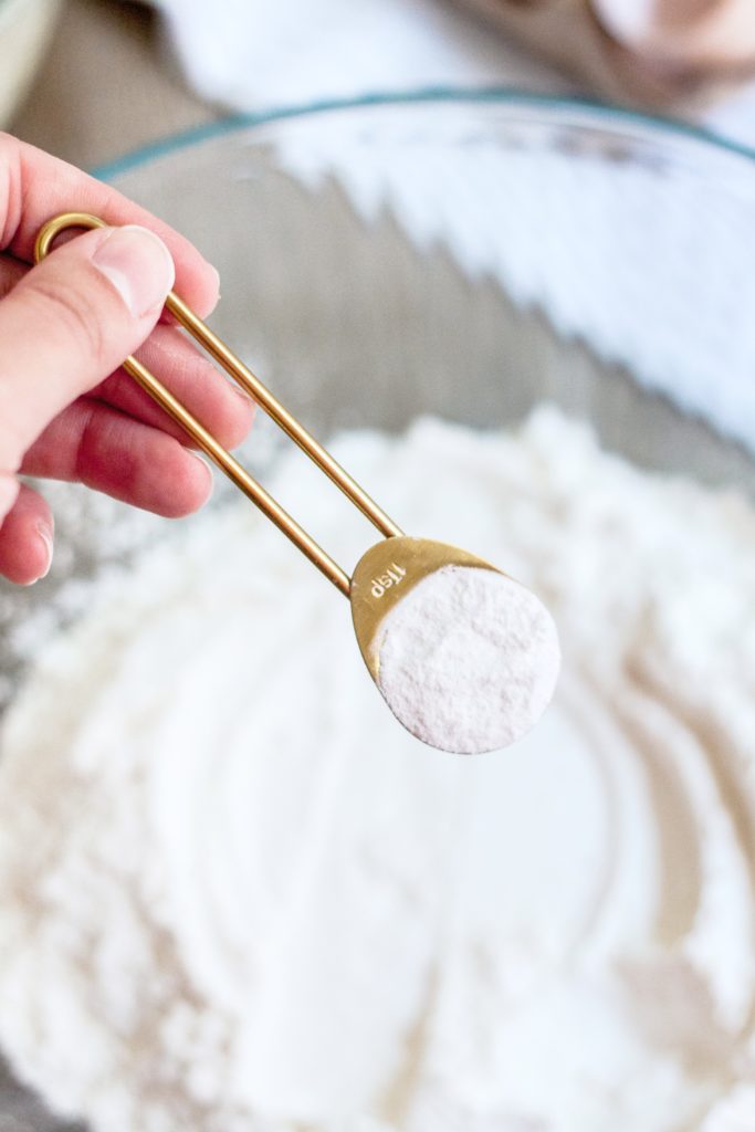 A gold measuring spoon is pouring baking powder onto flour.