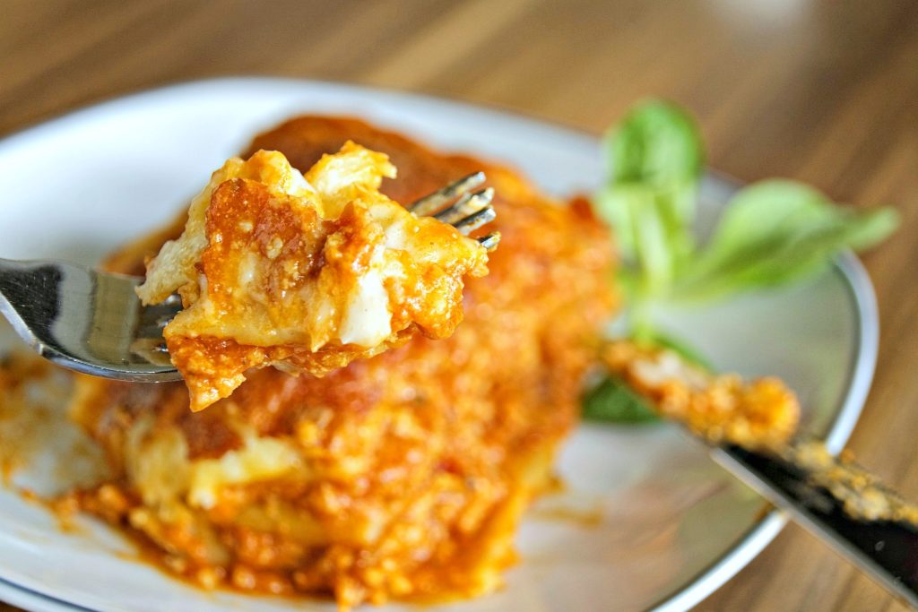 A fork is picking up a bite of lasagna and a piece of lasagna is shown.