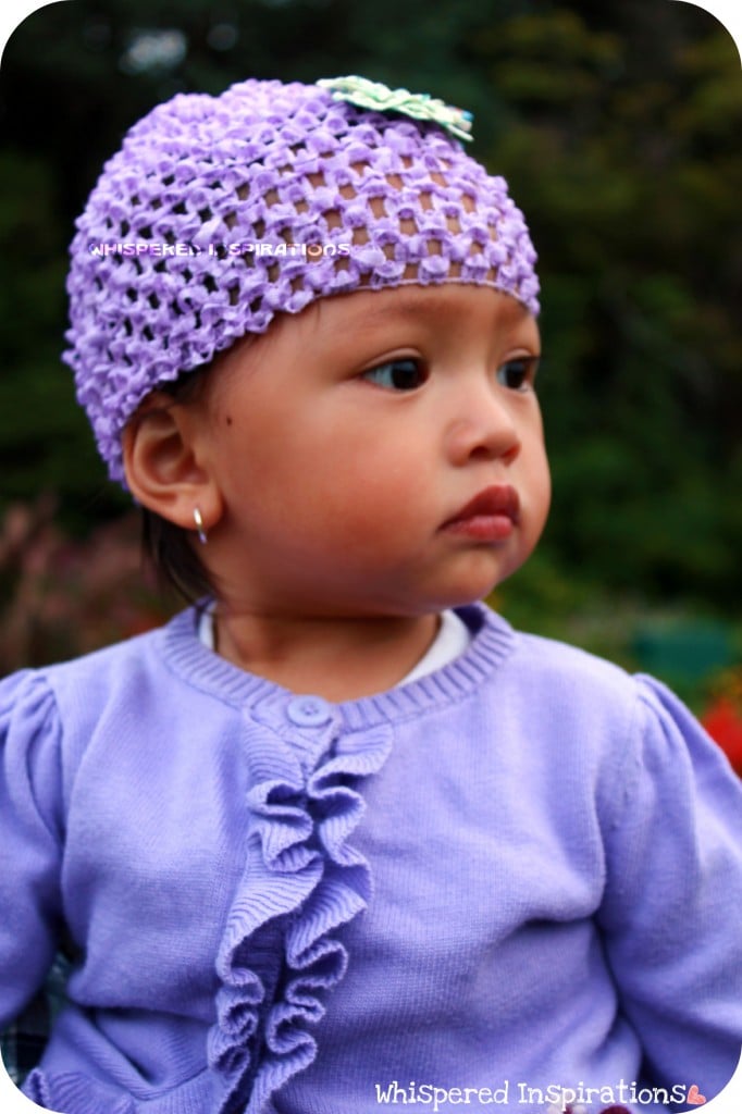 A baby is dressed in purple and is looking to her side, an earring is seen in her ear.