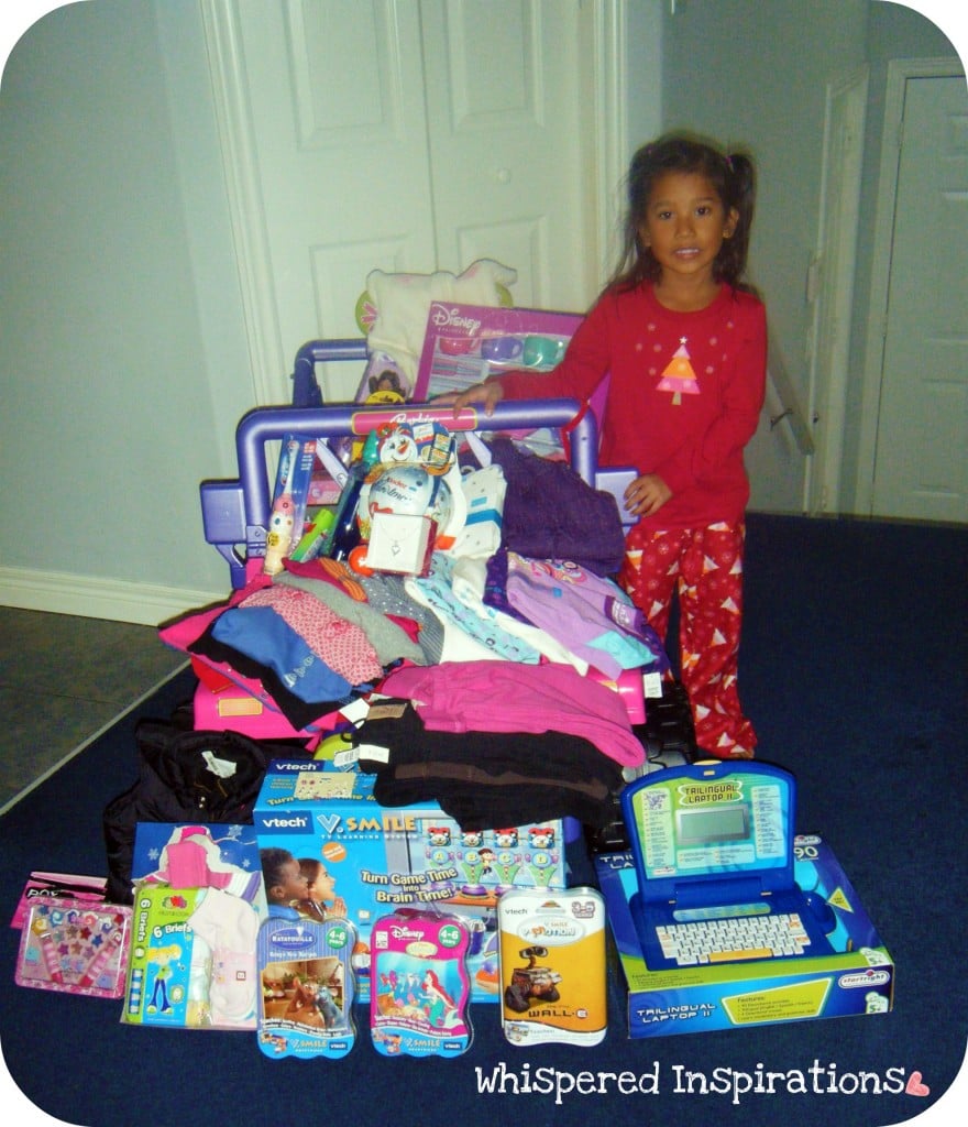 A little girl stands in front of toy Jeep and it is filled with gifts and she is surrounded by gifts.