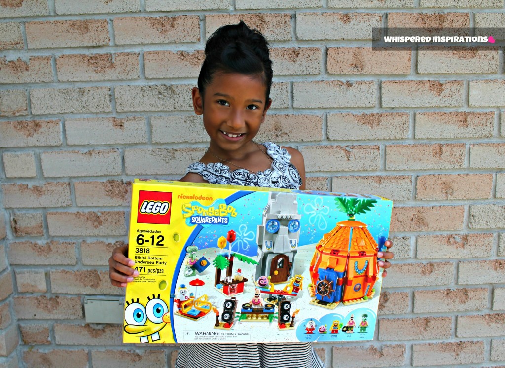 Little girl stands in front of a brick wall holding a large LEGO Spongebob Squarepants set.