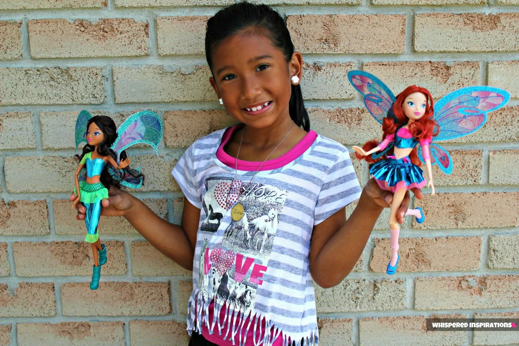 Gabby smiling against a brick wall and holding two Winx dolls. 