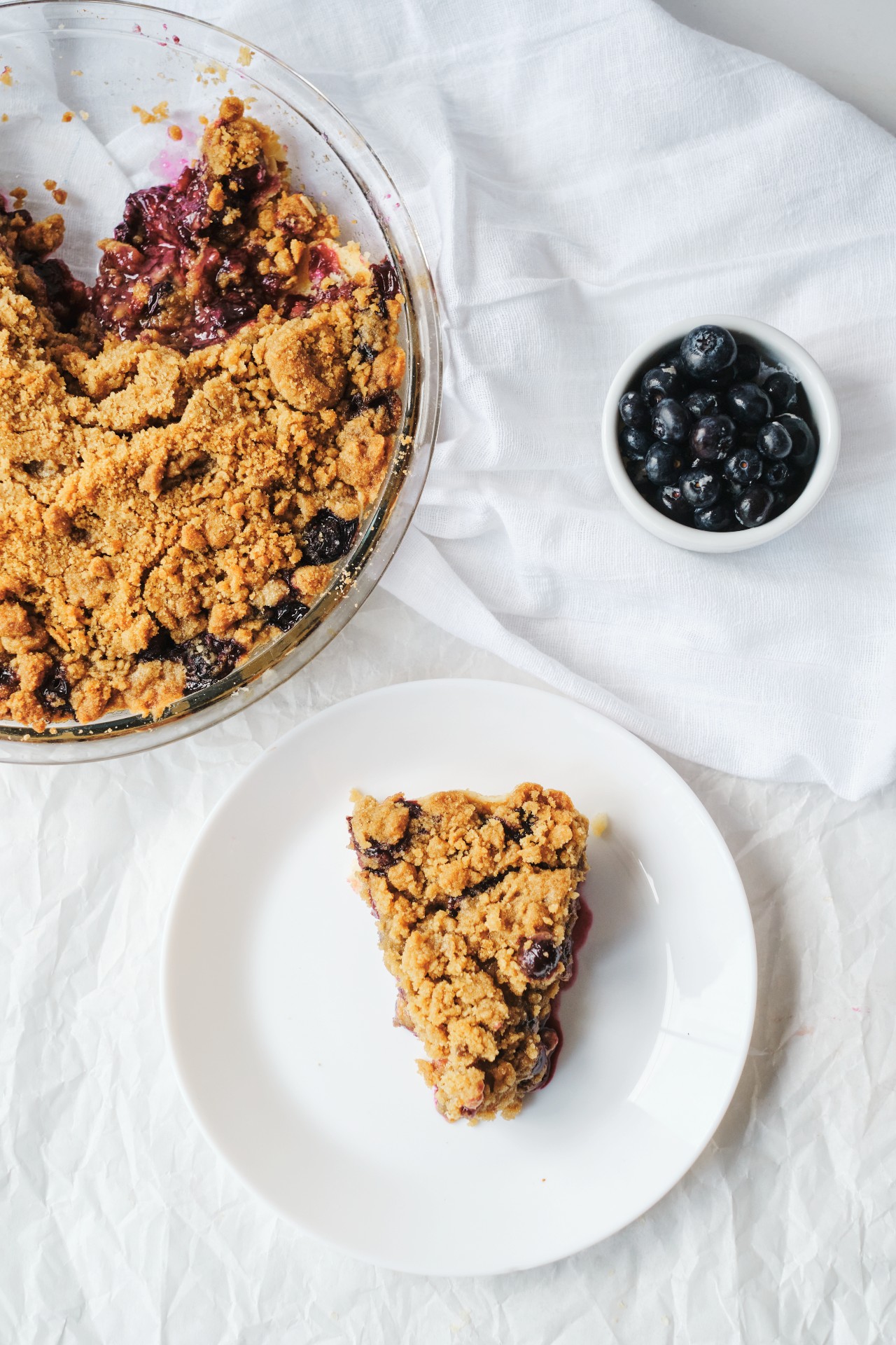 A blueberry crumble pie with a piece missing. The piece of pie is on a white plate on top of a napkin. There is a small bowl of blueberries shown. This article covers a Blueberry Crumble Pie recipe.