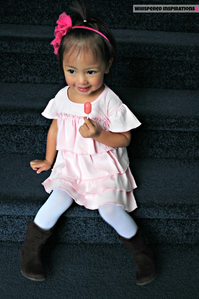 Little girl wears a pale pink organic dress with tights and boots while holding a lollipop.