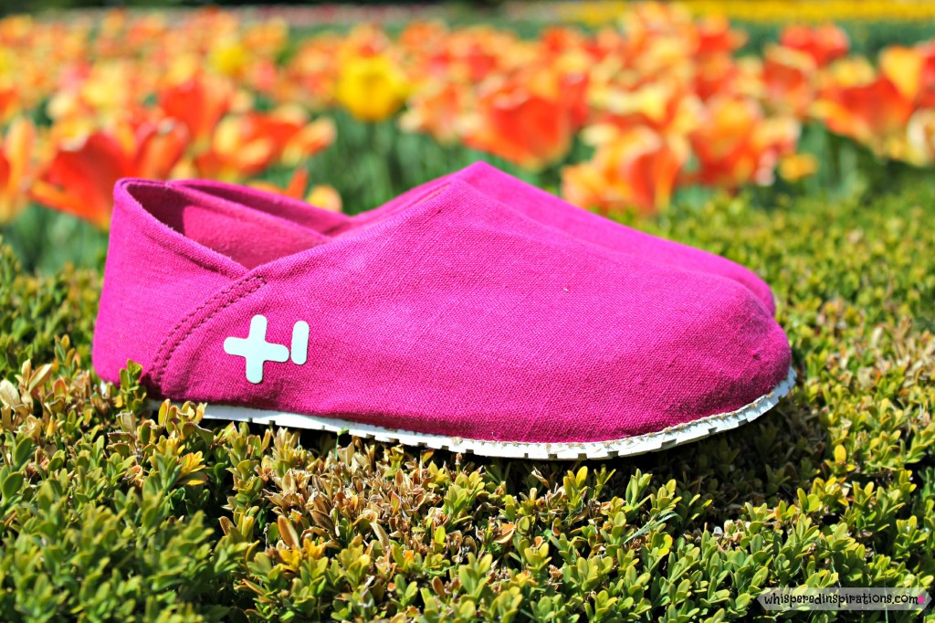 Hot pink pair of OTZ Shoes sit on a grassy surface with bright orange tulips behind it.