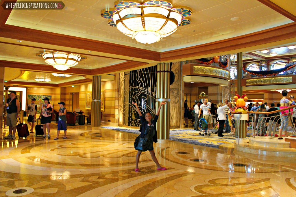 A little girl stands in the Disney Dream and holds her hands up in the air. The lobby is visible in the background. 