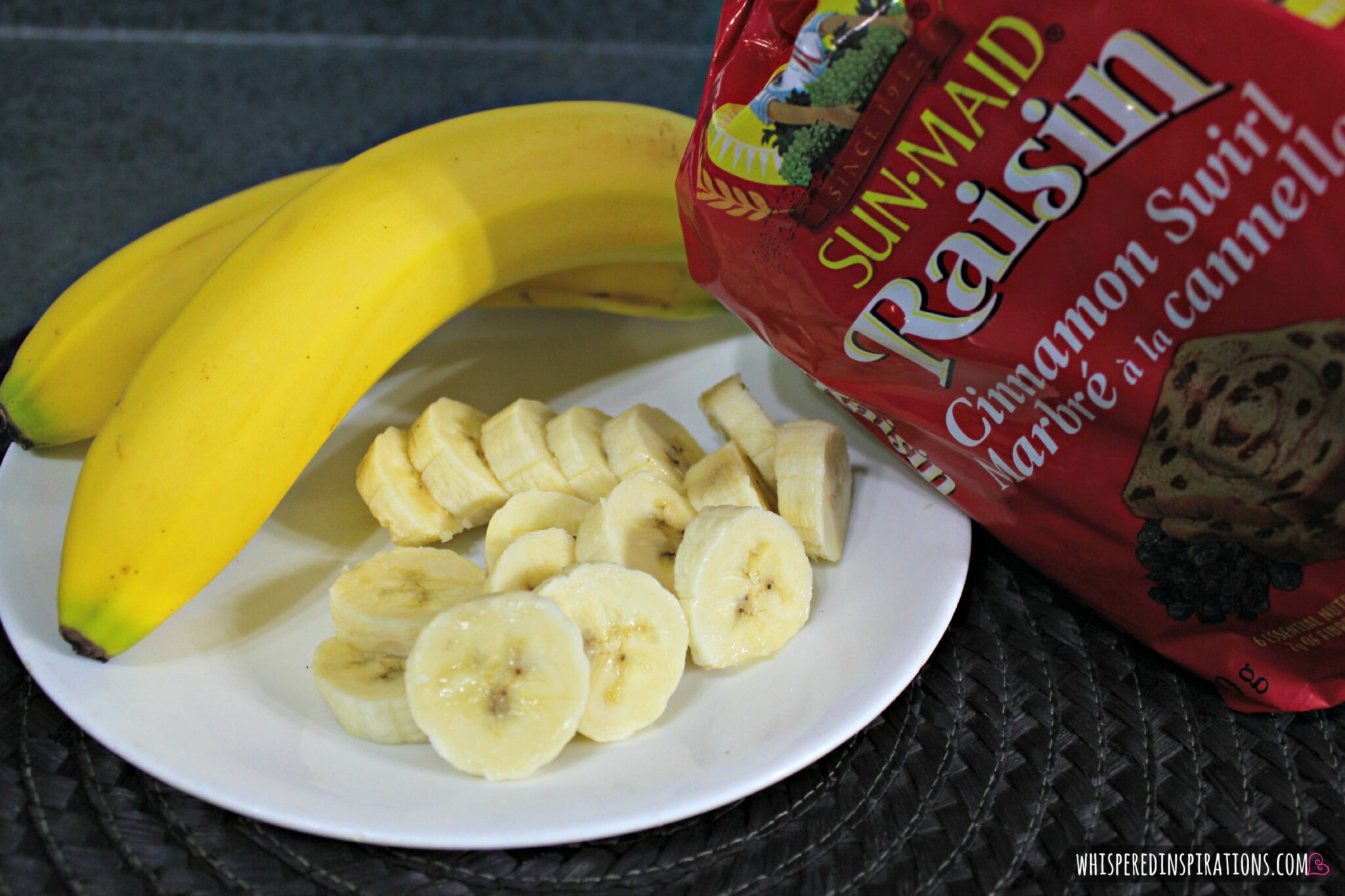 Sliced bananas on white plate with two fresh bananas and a loaf of Sun-Maid raisin bread.