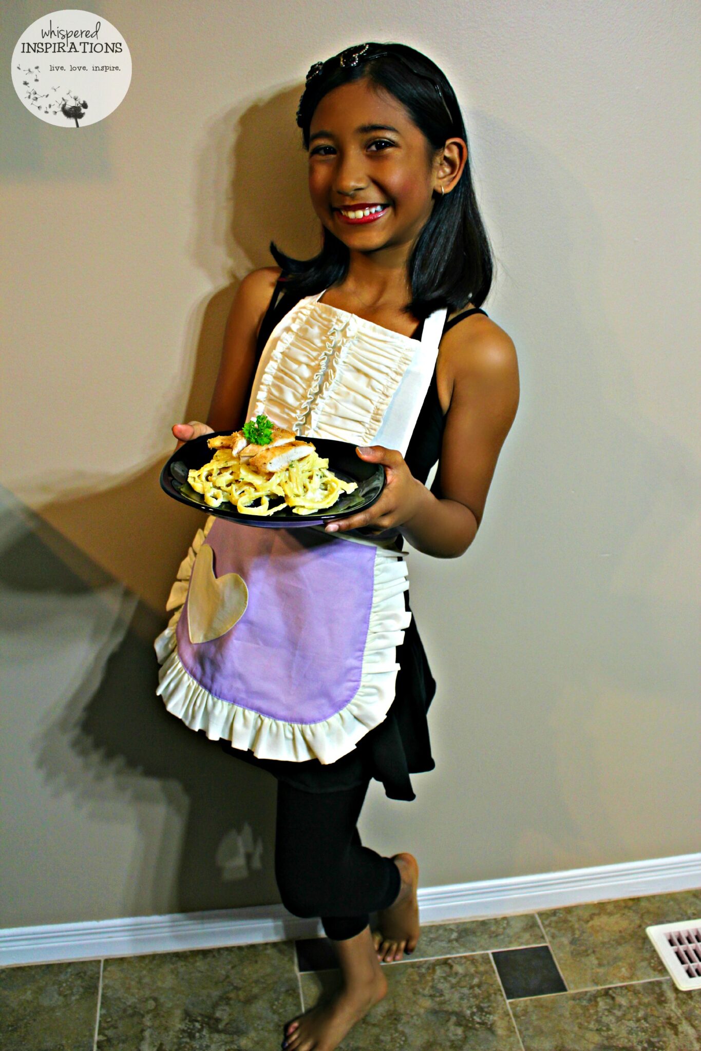 A little girl wears a Sugar Apron while holding the plate of Kids Chicken Fettuccine Alfredo pasta she just made. 