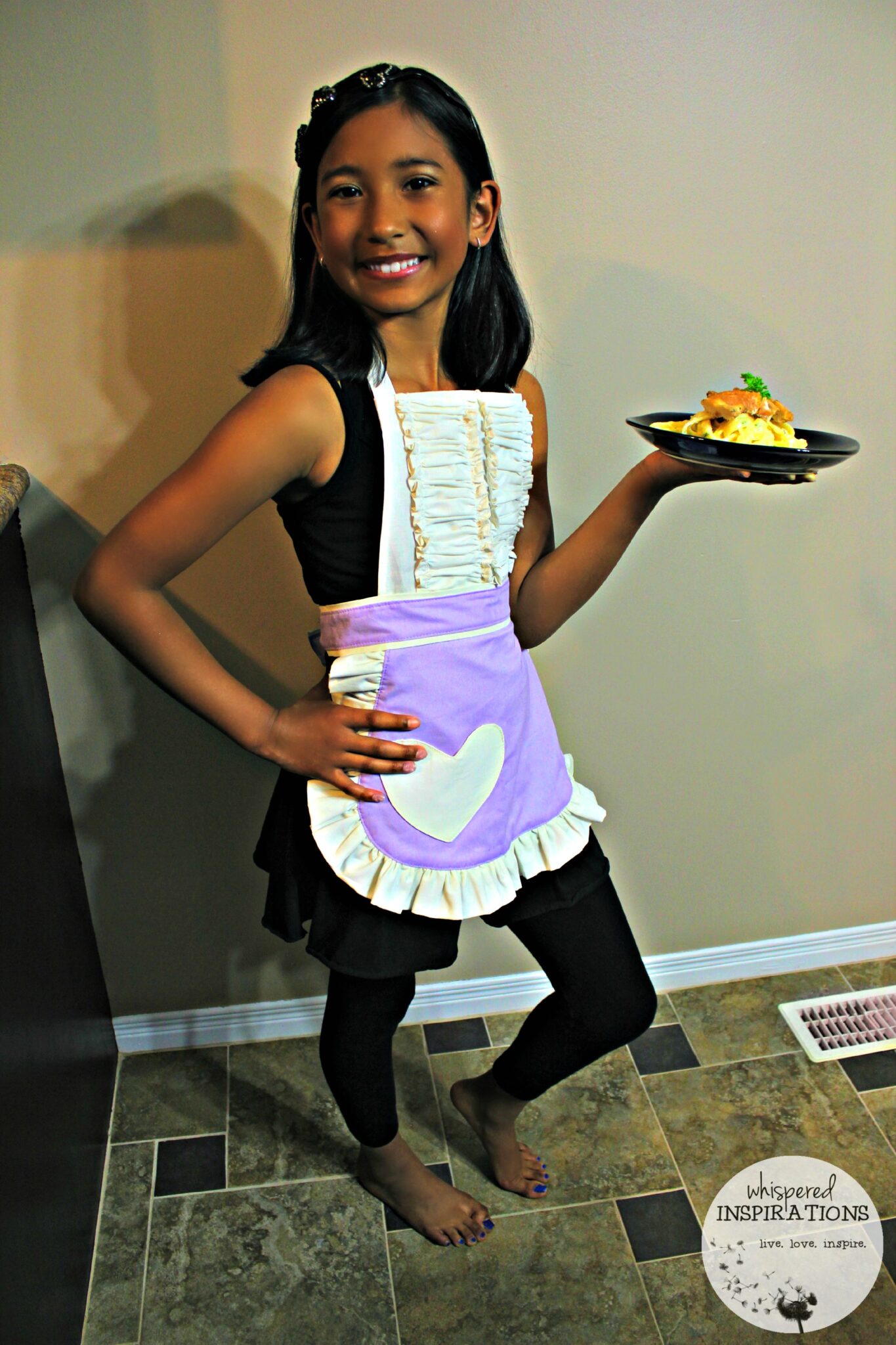 A little girl holds her plate of the Kids Chicken Fettuccine Alfredo and shows off her apron.