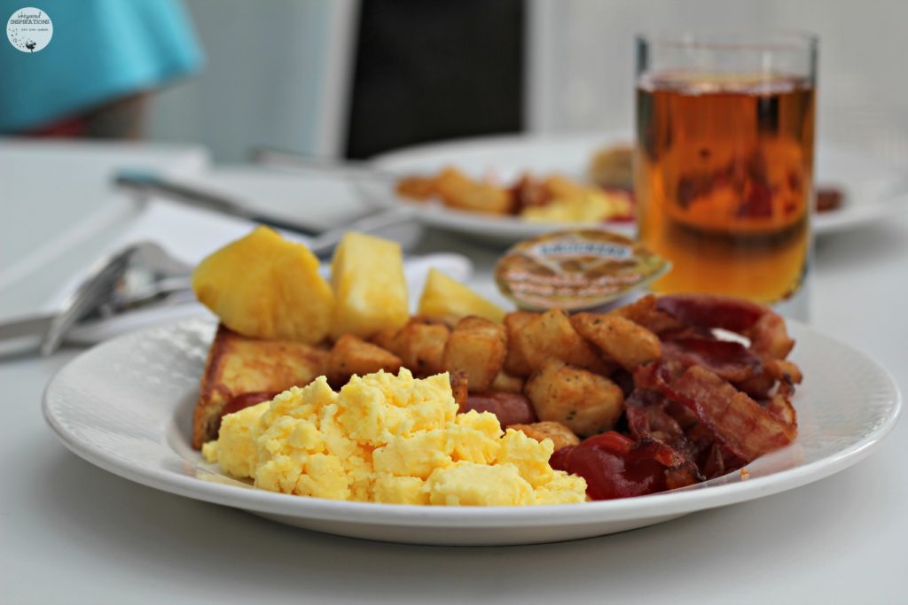 Breakfast plate: scrambled eggs, bacon, home fries, pineapple chunks, and apple juice. 