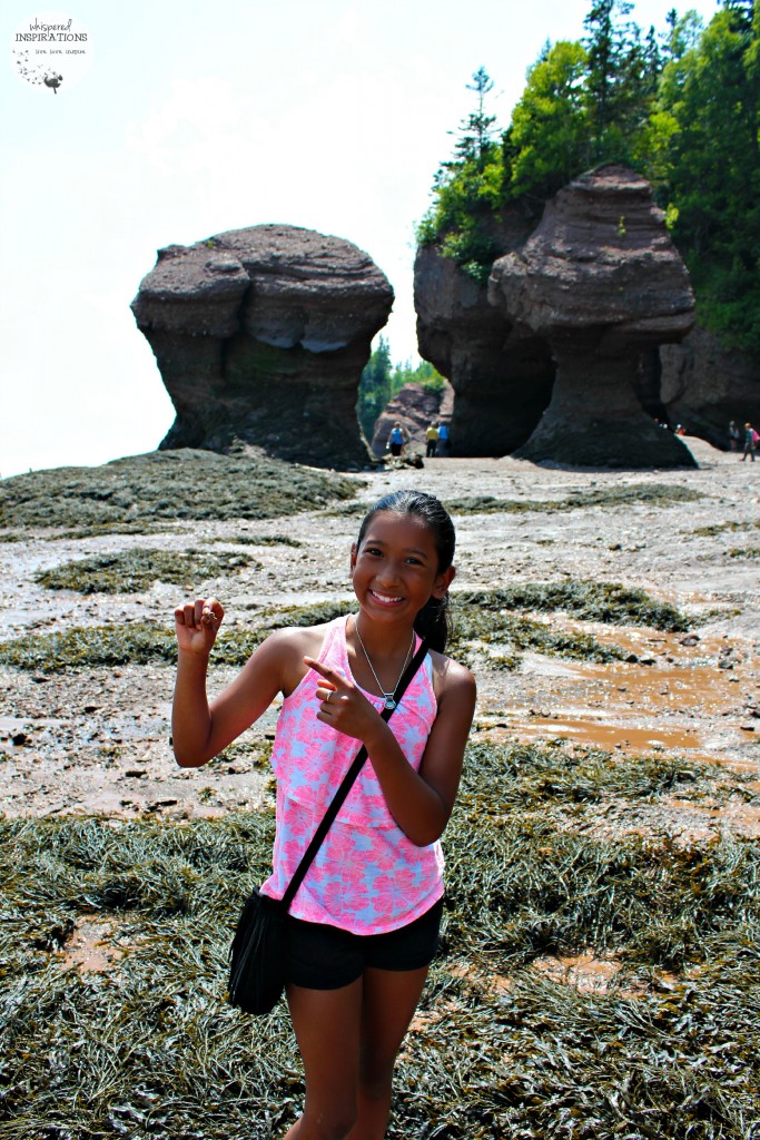 Young girl poses and points to a hermit crab in her hand. The Hopewell Rocks are in the distance.