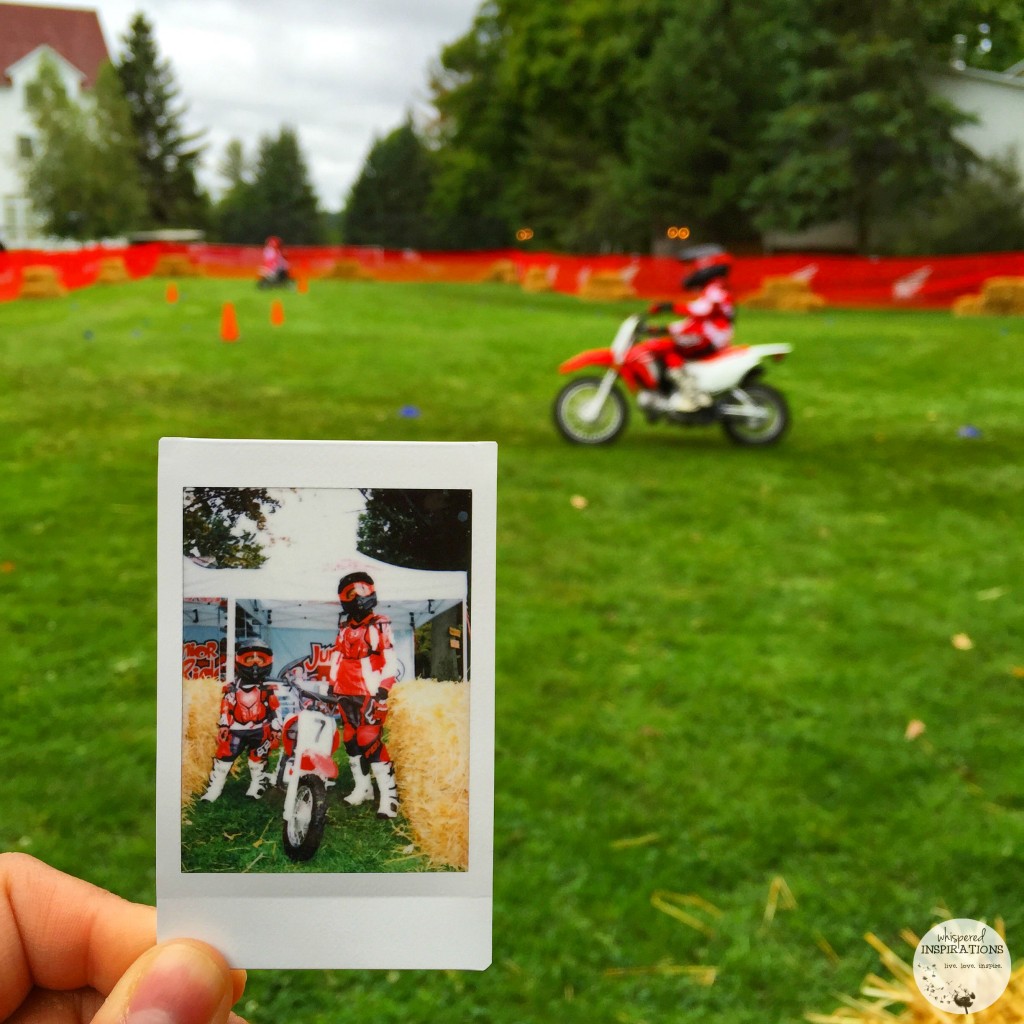 A Poloroid is held up against a picture of Gabby riding on a dirt bike. Gabby is blurred in the background riding the dirt bike. 