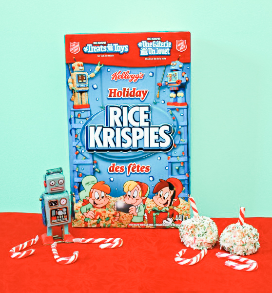 A box of Rice Krispies that are festive colors. Next to are candy canes, a toy robot, and the coconut white chocolate bite-sized rice treats.