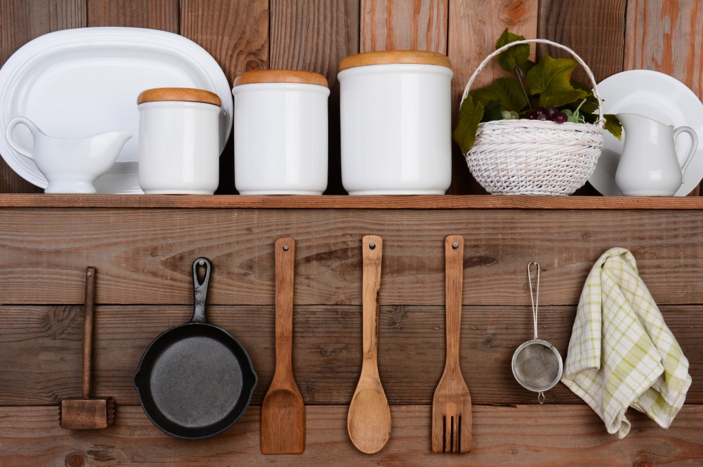 Closeup of a rustic kitchen wall. One shelf with canisters, plates and a basket. Hanging on the wall below are wooden utensils, frying pan and towel.