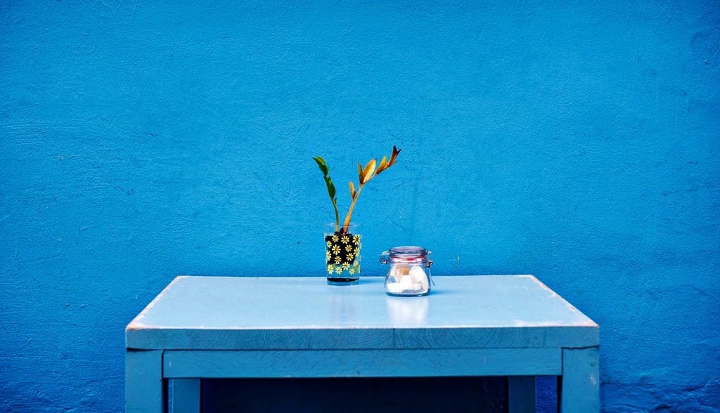 A bright blue wall with a blue table and plant and jar. 