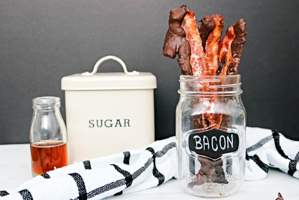 A mason jar holds chocolate-covered bacon and sugar and maple syrup and a striped napkin are shown. 