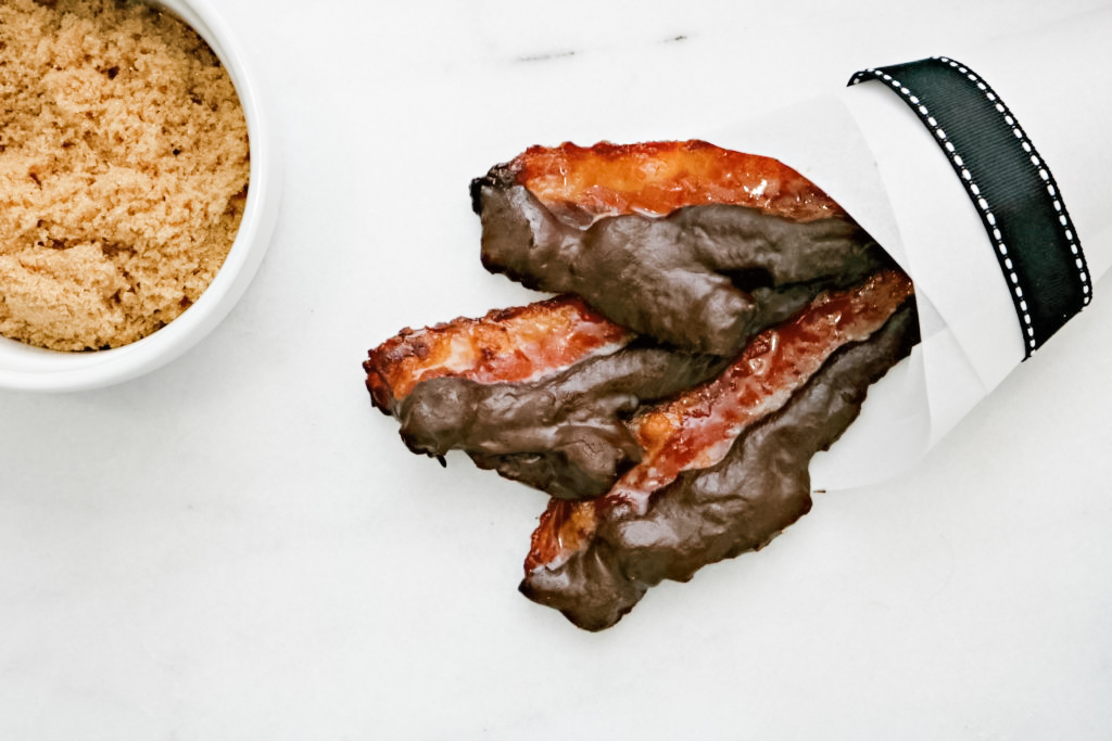 Close up of chocolate covered bacon and maple sugar next to it. Bacon is wrapped in paper and ribbon.