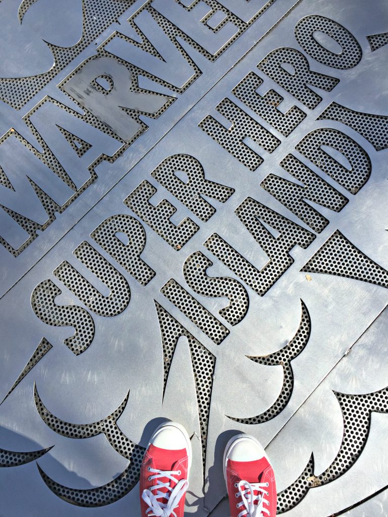 A woman stands on top of the Marvel Super Hero Island sign, her red Converse shoes are shown. 