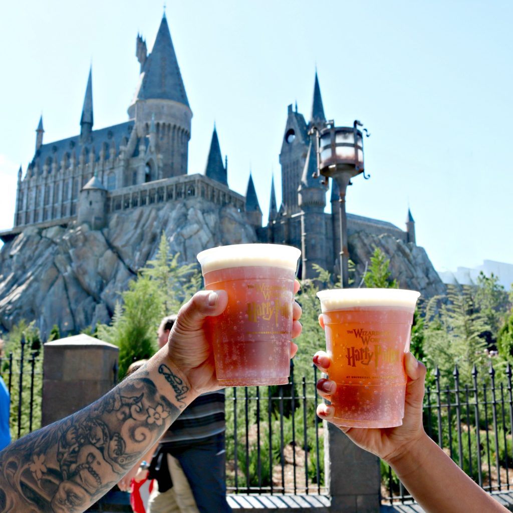 Two people hold butterbeer and do cheers in front of Hogwarts Castle. 