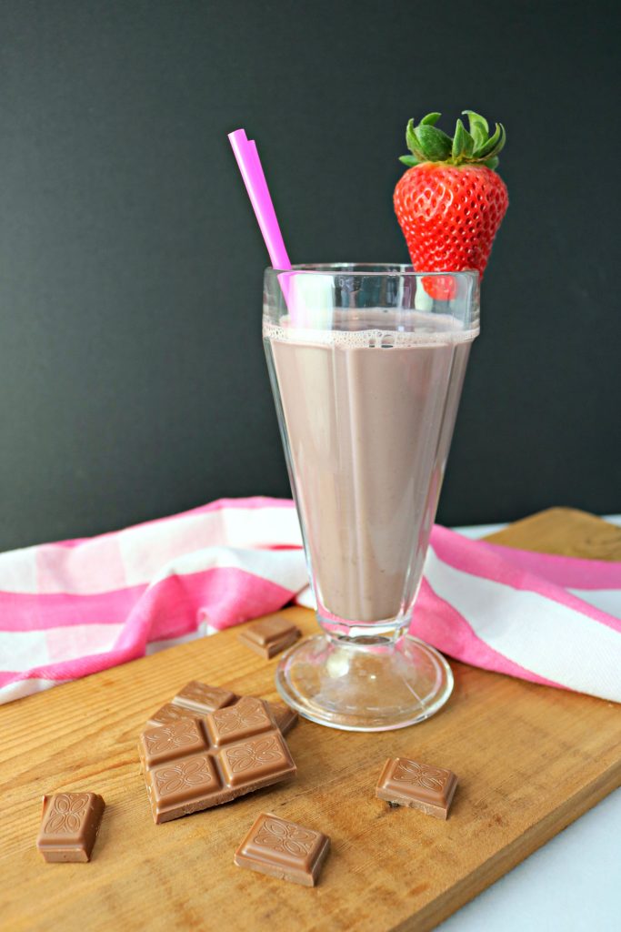 Milkshake glass on a board with napkin, straw, strawberry garnish, and chocolate pieces surrounding it. 