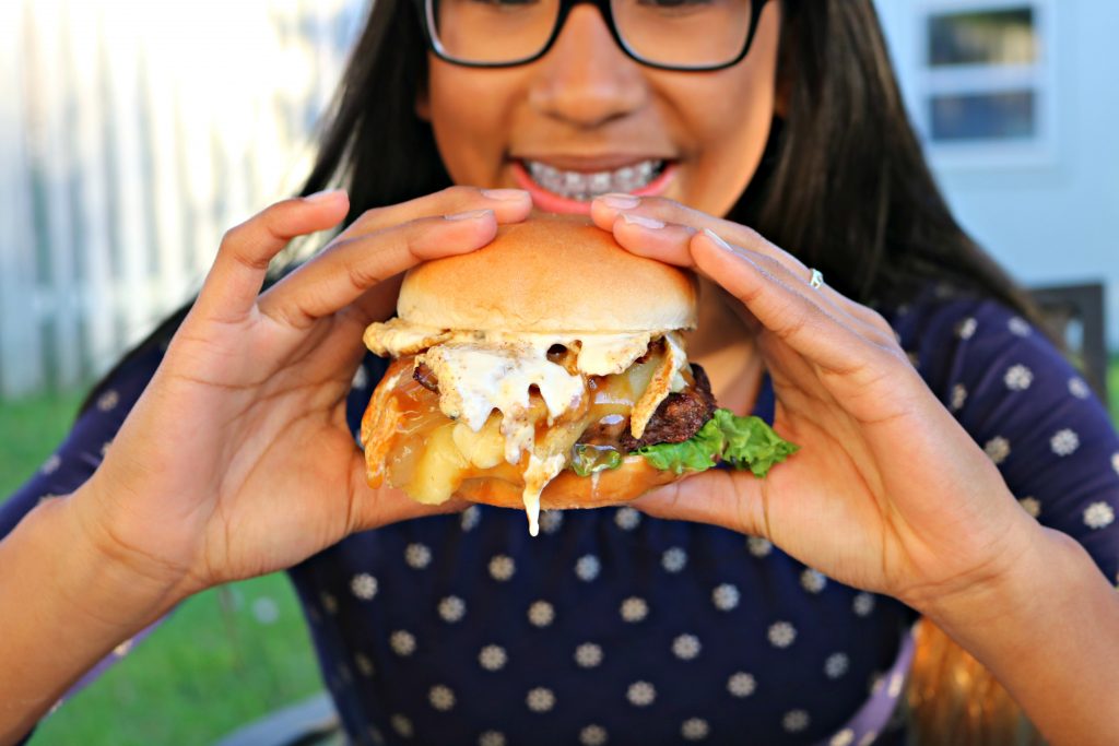 Gabby holds the burger as she takes a bite. The focus is on the burger, she is blurred. 