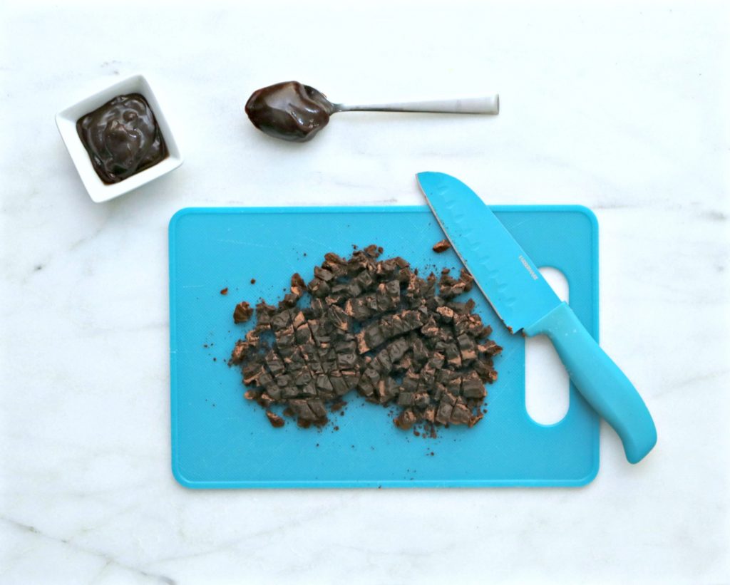 Blue cutting board with knife and cookies chopped coarsely. Chocolate sauce in a ramekin and on a spoon. 