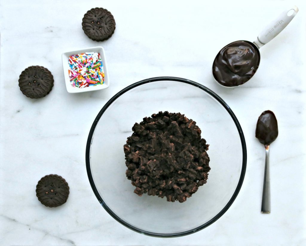 Clear bowl with chopped cookies, surrounded by 3 cookies, chocolate sauce in cup and spoon and a ramekin of sprinkles. 