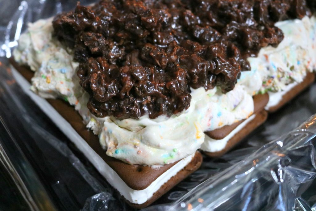 Side view of cake being layered with ice cream sandwiches, cookie and fudge mixture, and birthday cake ice cream. 
