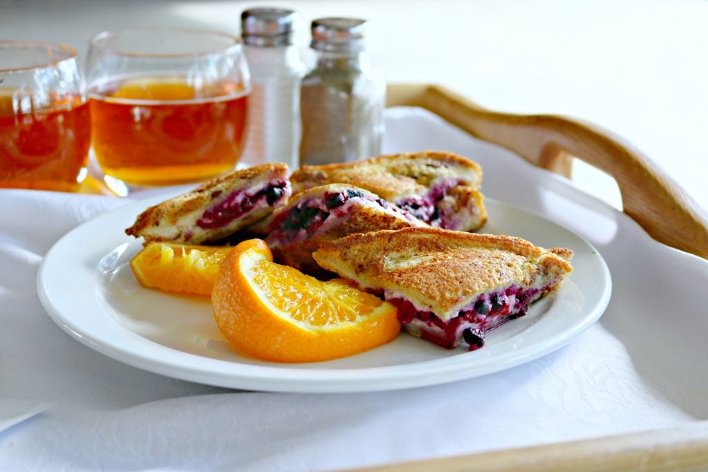 A delicious french toast filled with cream cheese and blueberries with a side of oranges on a plate. On a breakfast plate with salt and pepper and 2 glasses of apple juice. 