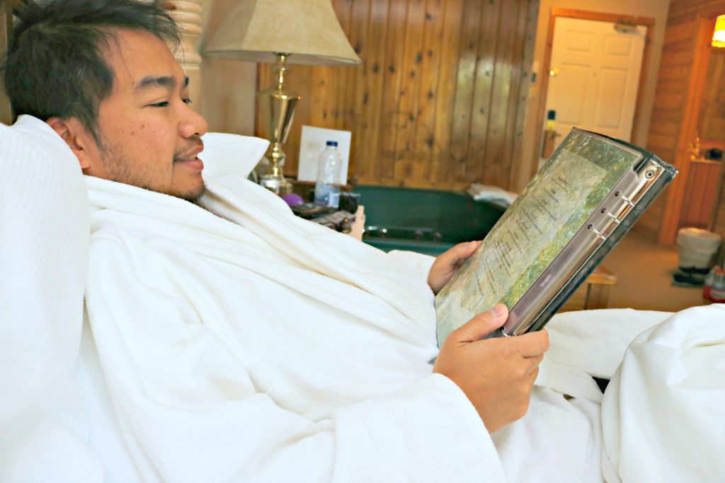 Darasak is in a robe in bed and looks at the room service menu. 