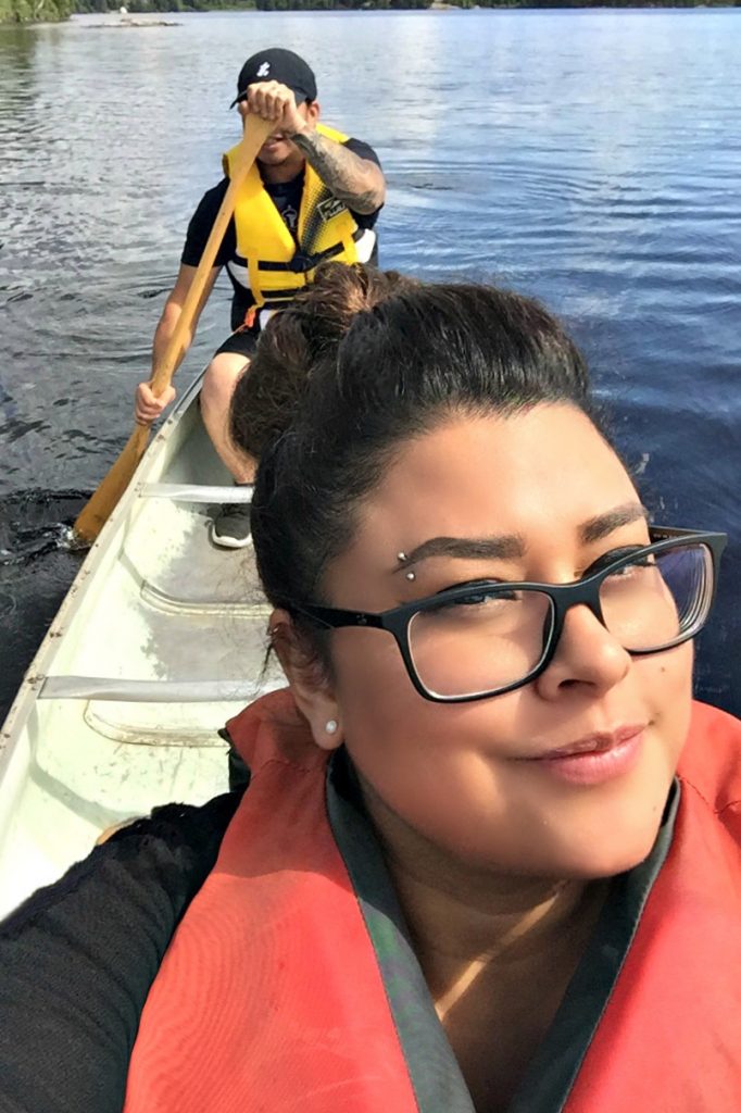 A wife and husband canoe on the lake, the wife takes a selfie and the husband paddles. 