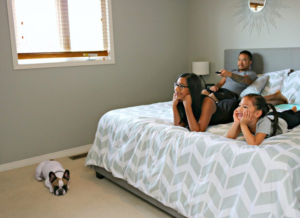A dad and his two daughters watch TV in his room. The family dog takes a nap on the carpet. 