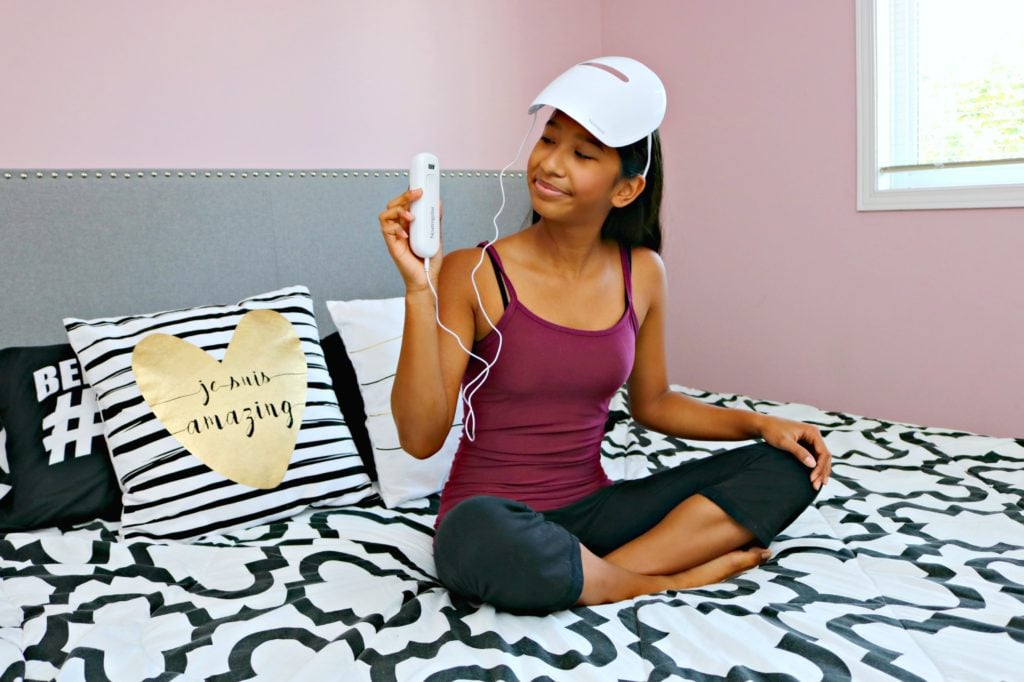 A teenager is holding the control to the Neutrogena Light Mask as she wears it on her head and sits on her bed.