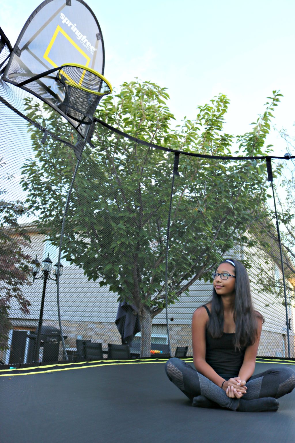 Girl sits pensively inside trampoline, basketball net can be seen and a backyard is behind her. Here are trampoline health benefits you never knew.