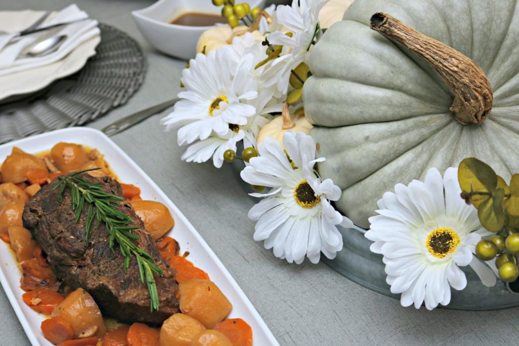 A close up of a beef roast with potatoes and carrots. A pumpkin centerpiece is shown.