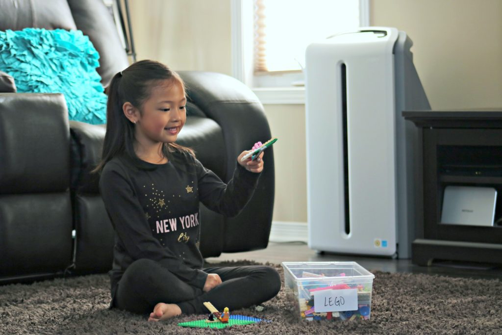 A little girl plays on her carpet in the living room with LEGOS, you can see the Atmosphere SKY Air Treatment machine behind her. 