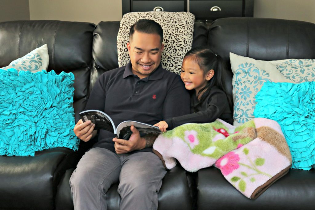 A father sits with his daughter on a couch and read a book.