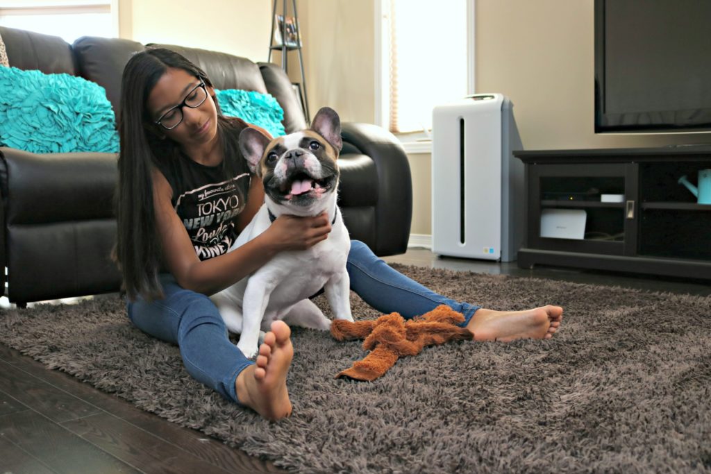 Gabby, a teen, sits and pets her dog on the carpet. The Atmosphere SKY Air Treatment System is seen in the back.