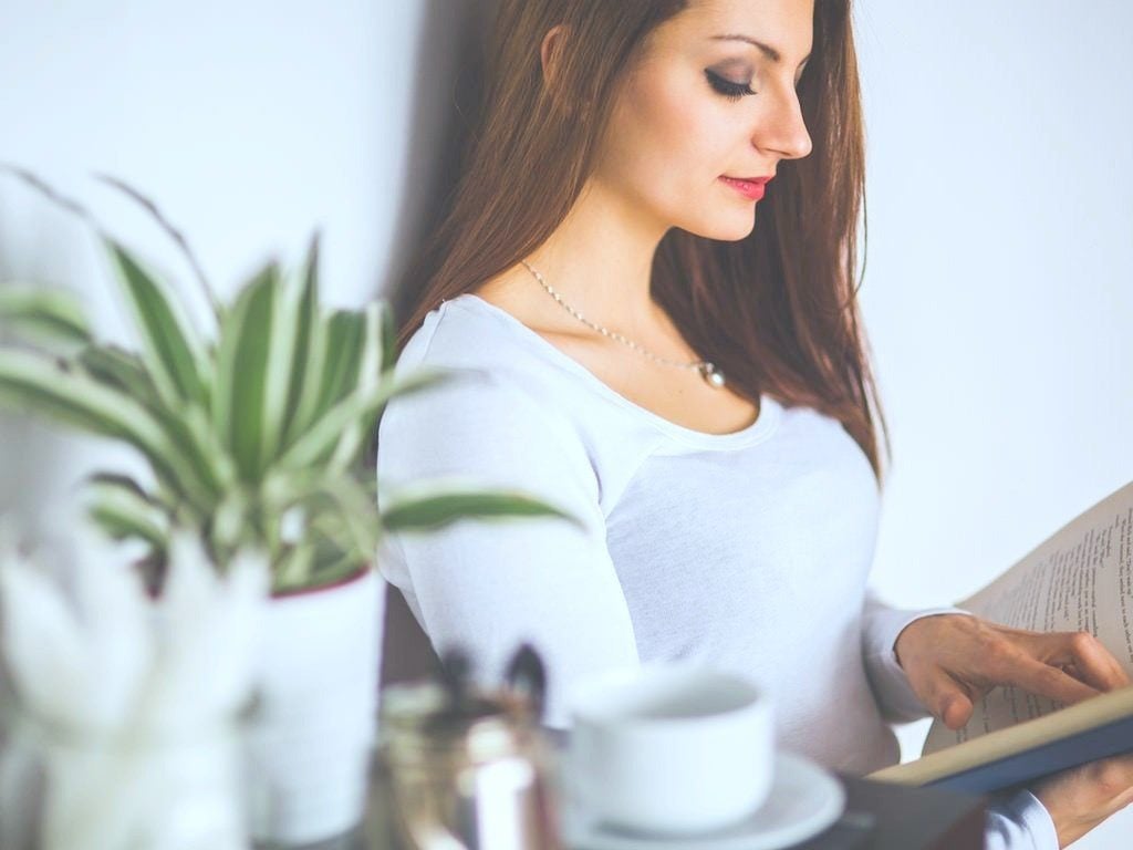 A woman leans against a wall, a table with a plant and tea are next to her. She is reading a book. This article covers how to learn to speak a foreign language from scratch.