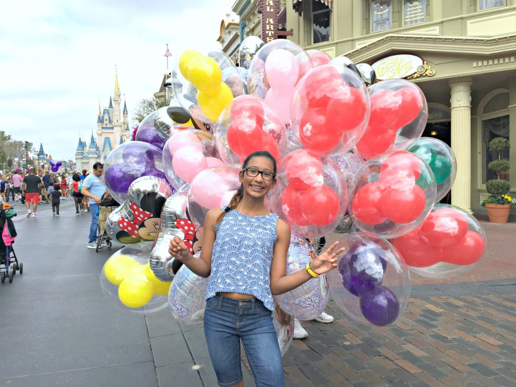 A happy girl stands in front of Mickey balloons at Magic Kingdom at Disney World.