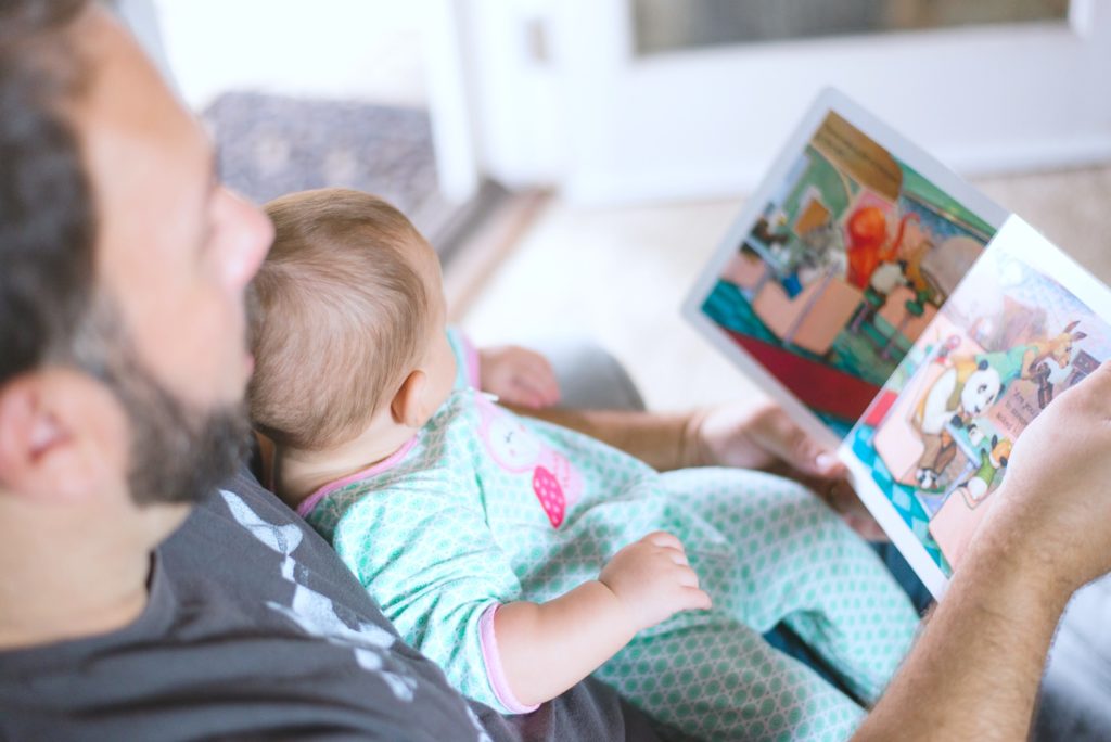 Baby sits and reads with her father on couch.