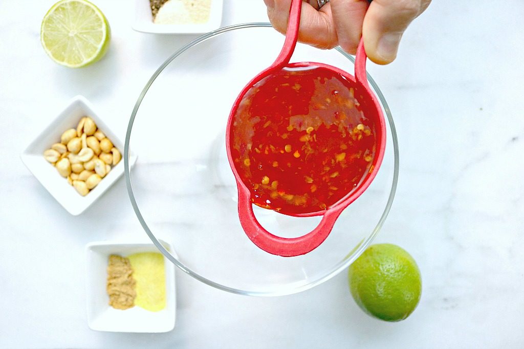 An empty bowl is shown surrounded by ingredients and a half cup of Thai chili sauce about to be poured in.