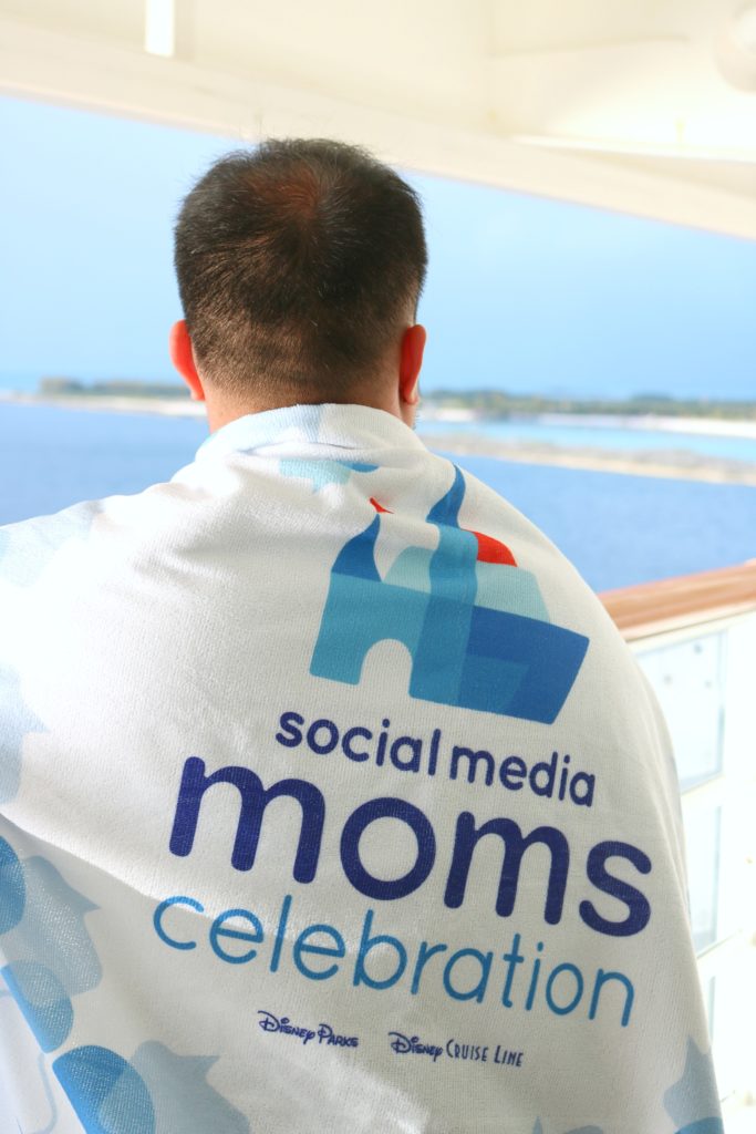 A man looks out to Castaway Cay with the Disney Social Media Moms Celebration towel on his back. 