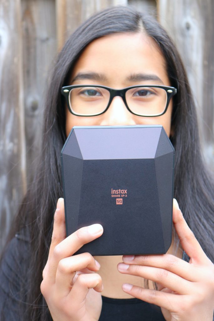 A girl smiles at the camera holding the Instax printer on half of her face playfully.