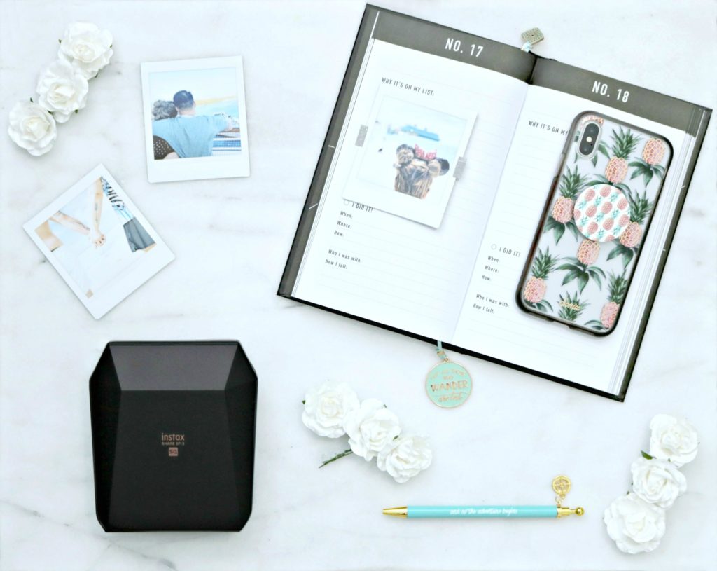 An open bucket list journal with Instax printer, printed out pictures, a pen, and a phone are shown.