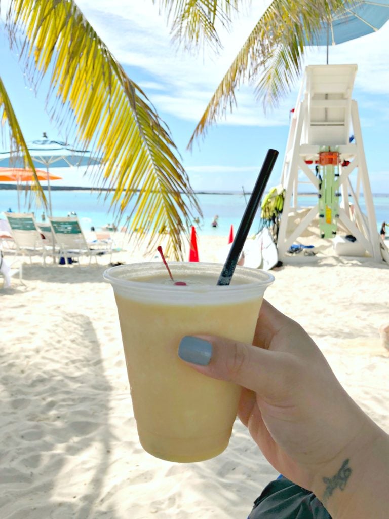 A palm tree frames a view of crystal clear blue waters while a woman holds up a drink to the camera.