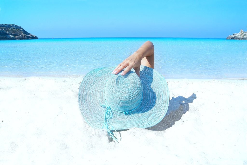 Woman lays on the beach while holding on to her blue beach hat. 