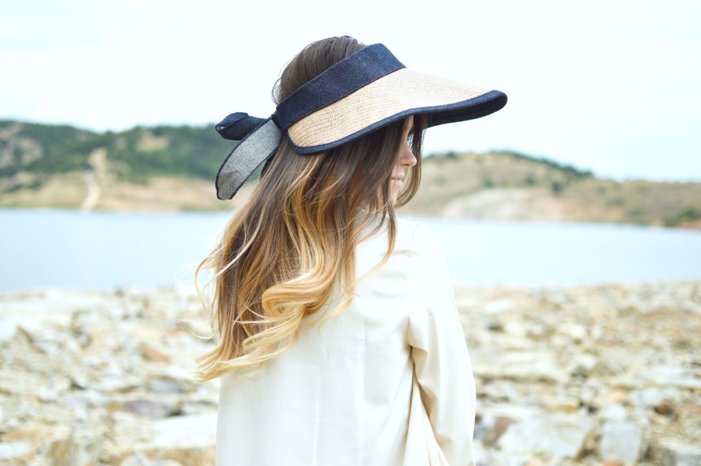 Woman sits by the water and looks to the side, hat hides her face.