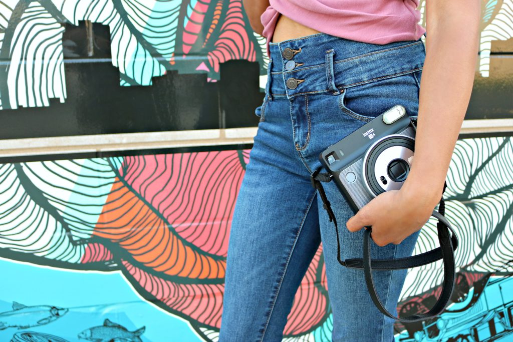 A girl holds a camera in front of a graffiti wall. 